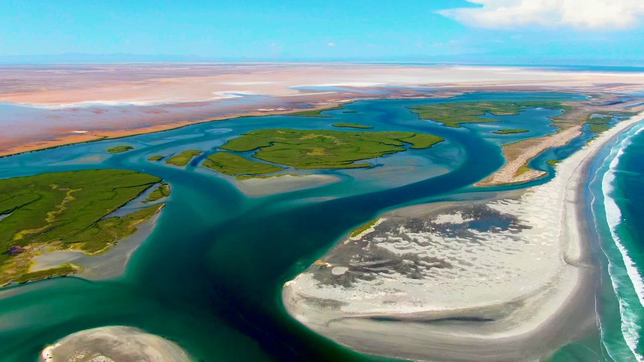 Aerial coastal channels winding around sandbars and green land under a blue sky.