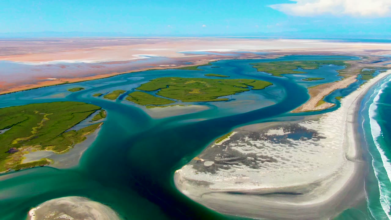 Aerial frame over La Bocana, Baja California Sur showing branching blue-green waterways, a bright sandbar in the lower right, surf breaking along the coast, and no visible people.