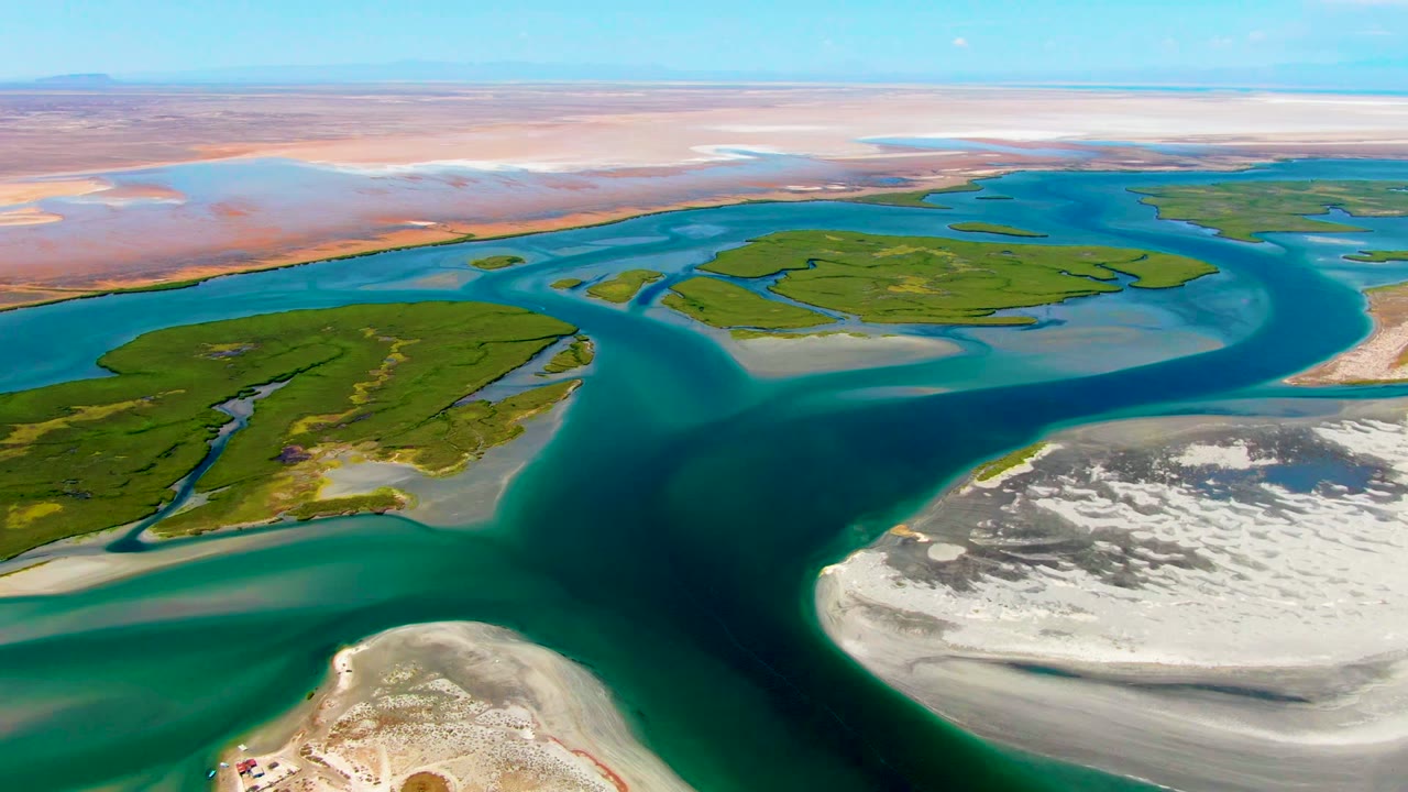 Opening aerial frame over La Bocana, Baja California Sur showing teal river channels around green islands, pale sandbars in the foreground, arid flats beyond, and no visible people.