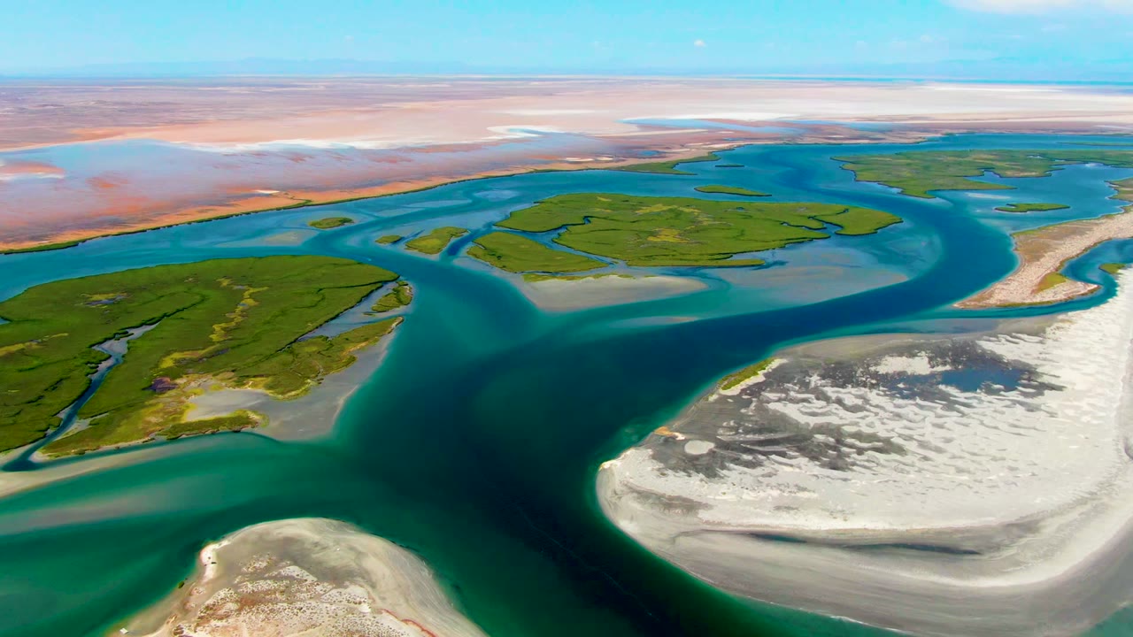 Opening aerial frame over La Bocana, Baja California Sur, with turquoise river channels wrapping green islands, white sandbars in the foreground, and no visible people.