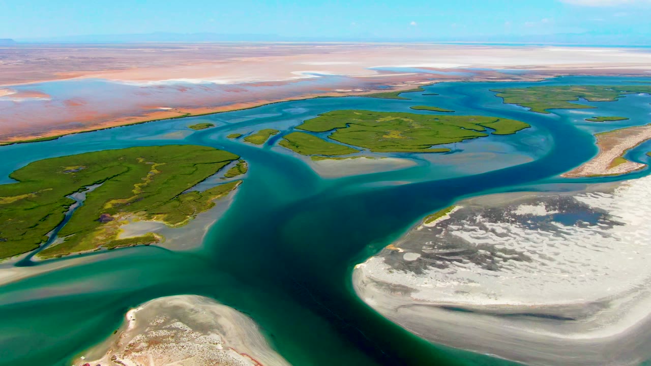 La Bocana delta aerial with turquoise water separating green island patches, a broad white sandbar at lower right, distant tan flats, and no visible people.