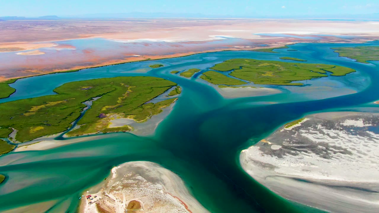 Drone view of blue water curving past green marsh islands and pale sand.