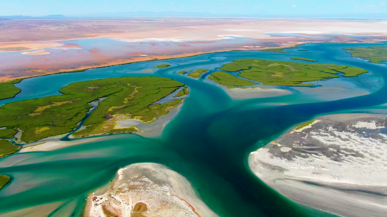 Aerial frame over La Bocana showing green island shapes divided by turquoise and dark blue water, pale sandbars at the lower edge and right side, pink-brown flats beyond, and no visible people.