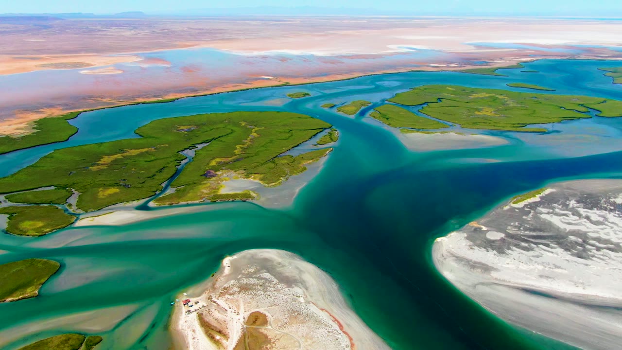 Aerial view over La Bocana, Baja California Sur, with turquoise delta water curving around green islands, pale sandbars in the foreground, and no visible people.