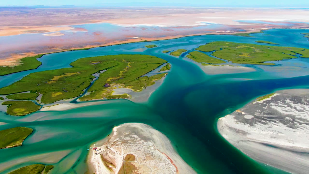Wide drone view of a delta with teal water, green marsh islands, and exposed sand.