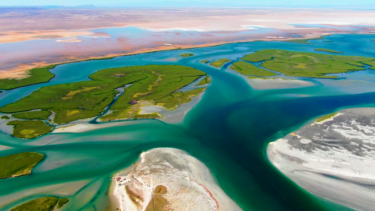 La Bocana aerial frame with a darker central channel bordered by turquoise shallows, green island edges, white sand flats, and no visible people.