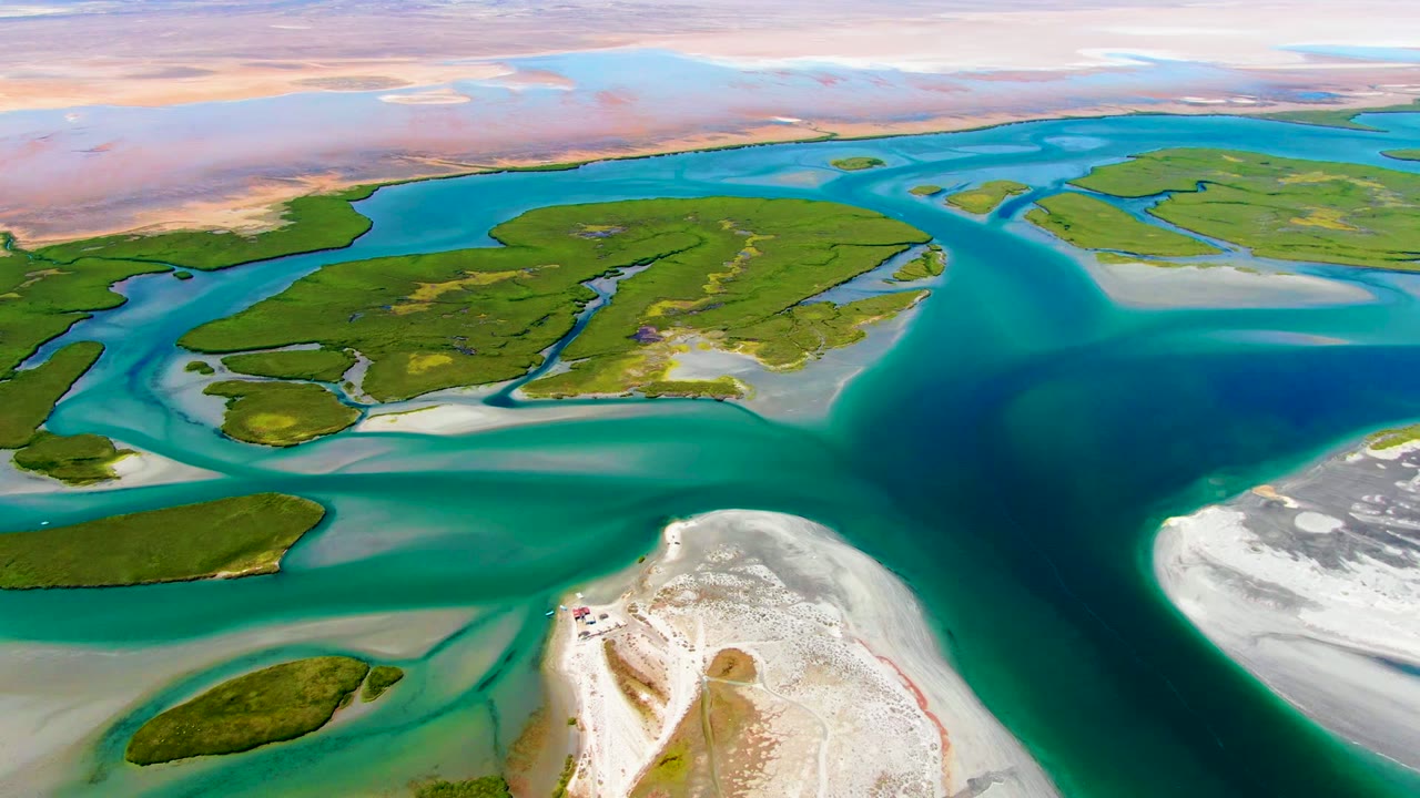 Opening aerial frame over La Bocana, Baja California Sur, showing green islands split by turquoise tidal channels, pale sand in the foreground, distant tan land, and no visible people.