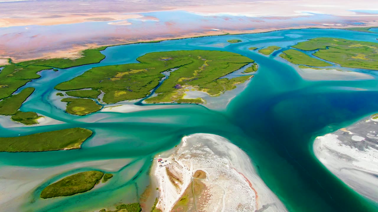 Mid-clip aerial view over La Bocana showing branching turquoise channels around green islands, pale sandy flats on the right and lower foreground, and no visible people.