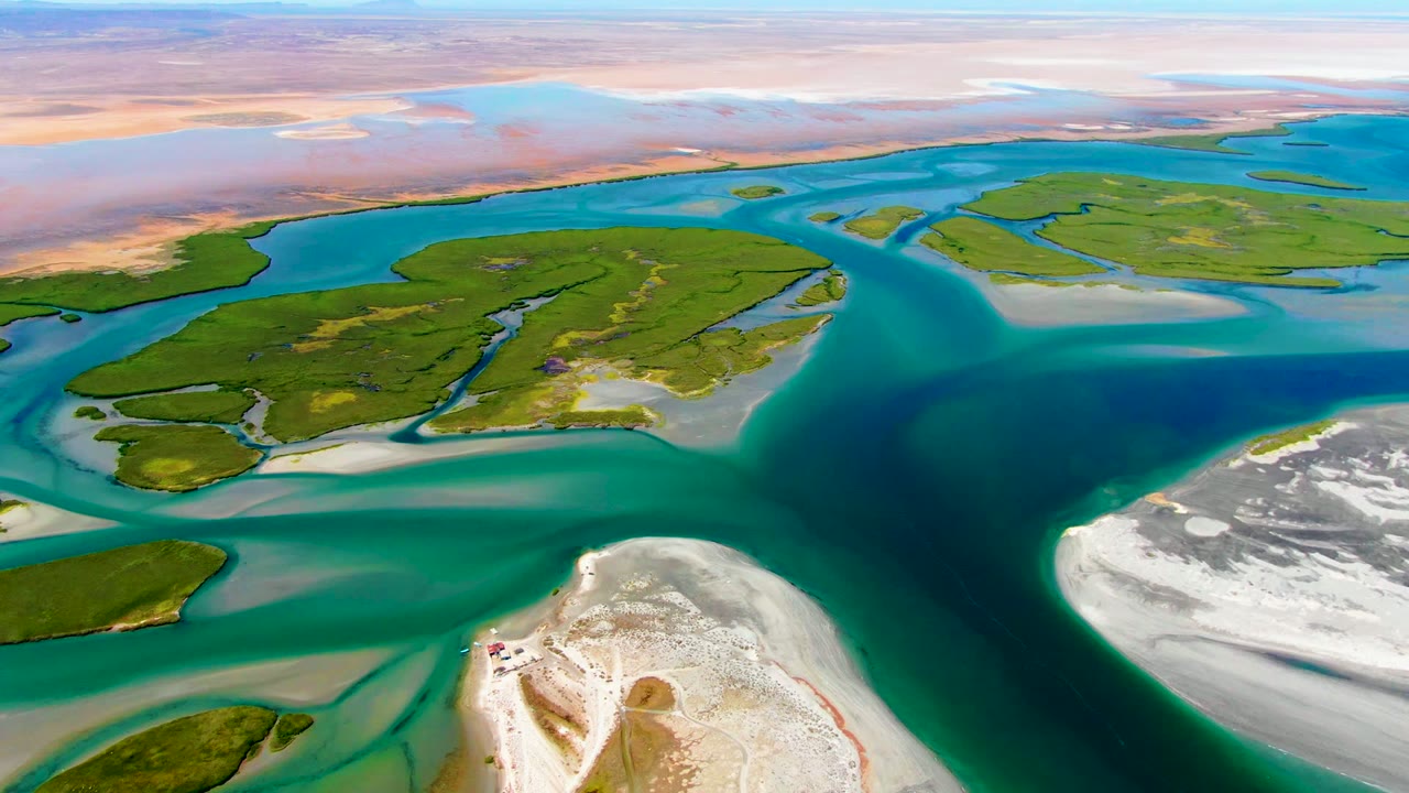 Opening aerial frame over La Bocana, Baja California Sur, with green delta islands divided by turquoise channels, pale sandbars, white shoreline in the foreground, and no visible people.