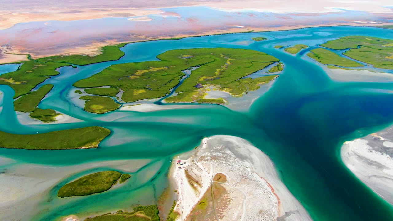 Opening aerial frame over La Bocana, Baja California Sur, with turquoise channels wrapping green islands, a white sandbar in the foreground, pinkish flats on the horizon, and no visible people.