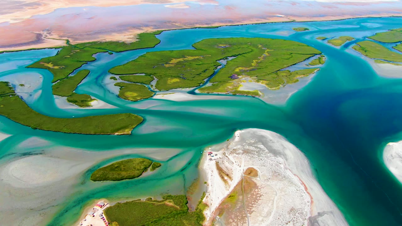 Opening aerial frame over La Bocana, Baja California Sur, showing green lagoon islands, turquoise shallow channels, pale sandbars, a deep blue inlet, and no visible people.