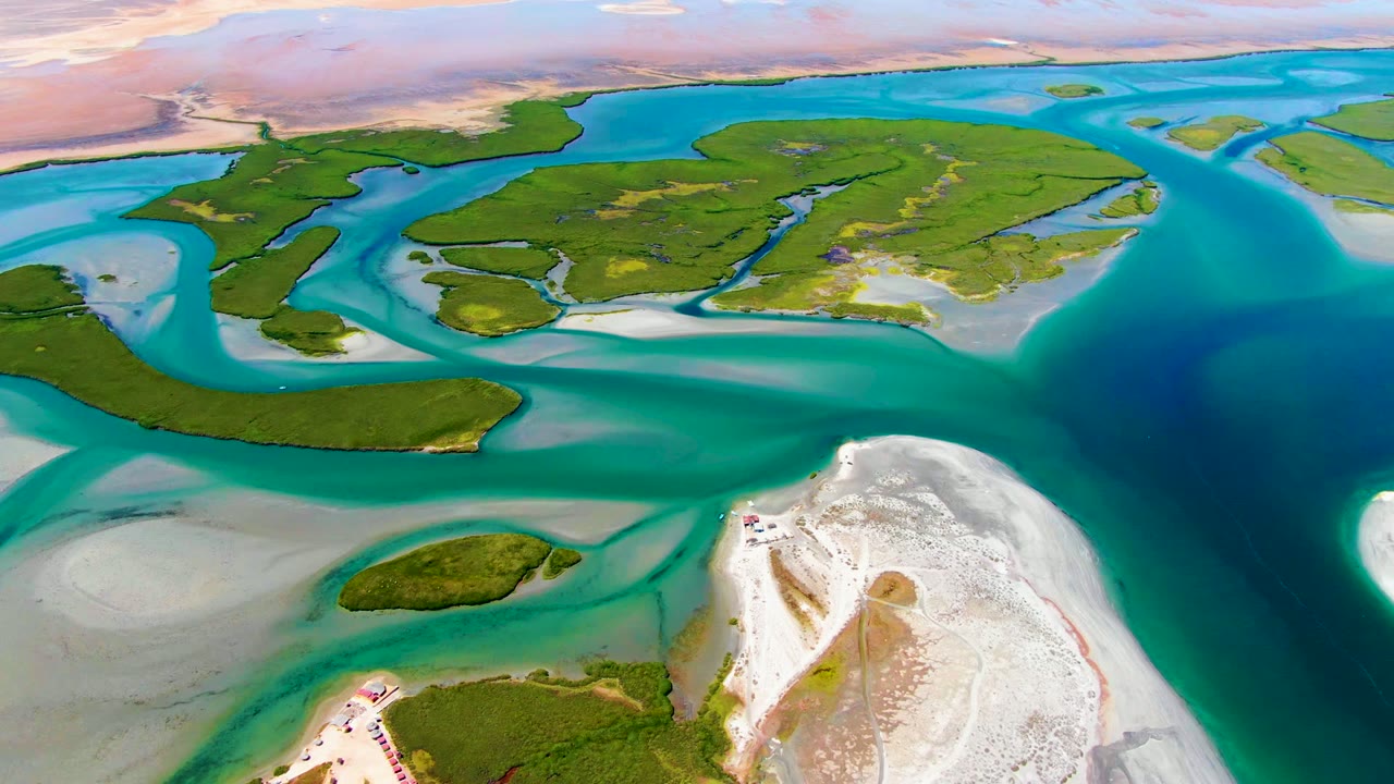 Mid-clip aerial view of La Bocana showing green island shapes across the upper frame, turquoise shallows, a dark blue water channel at right, white sand at lower right, and no visible people.