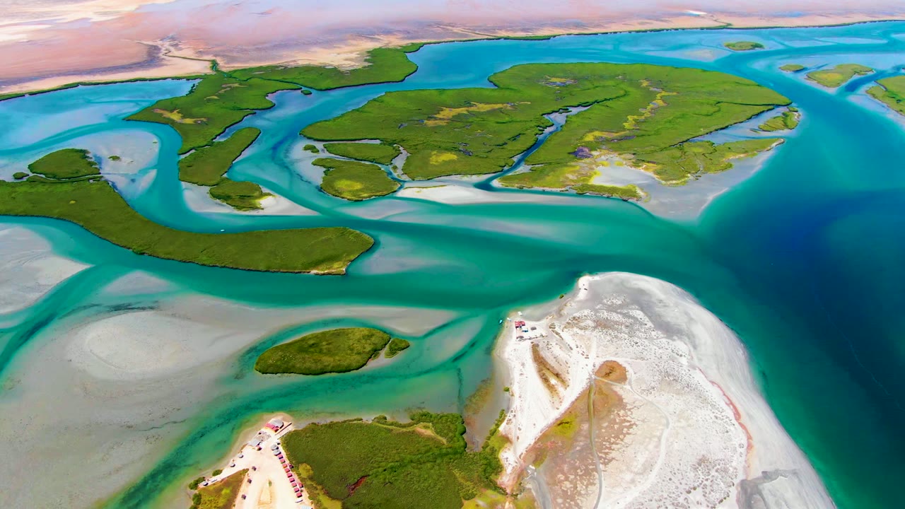 Aerial view over La Bocana, Baja California Sur, with turquoise channels wrapping green tidal islands, white sandbars in the foreground, distant arid land, and no visible people.