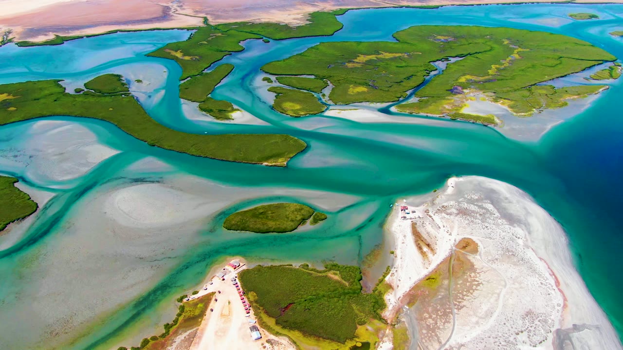 At 0:00, a high aerial view over La Bocana shows turquoise channels around green islands, pale tidal sand, a white shoreline, and no visible people.