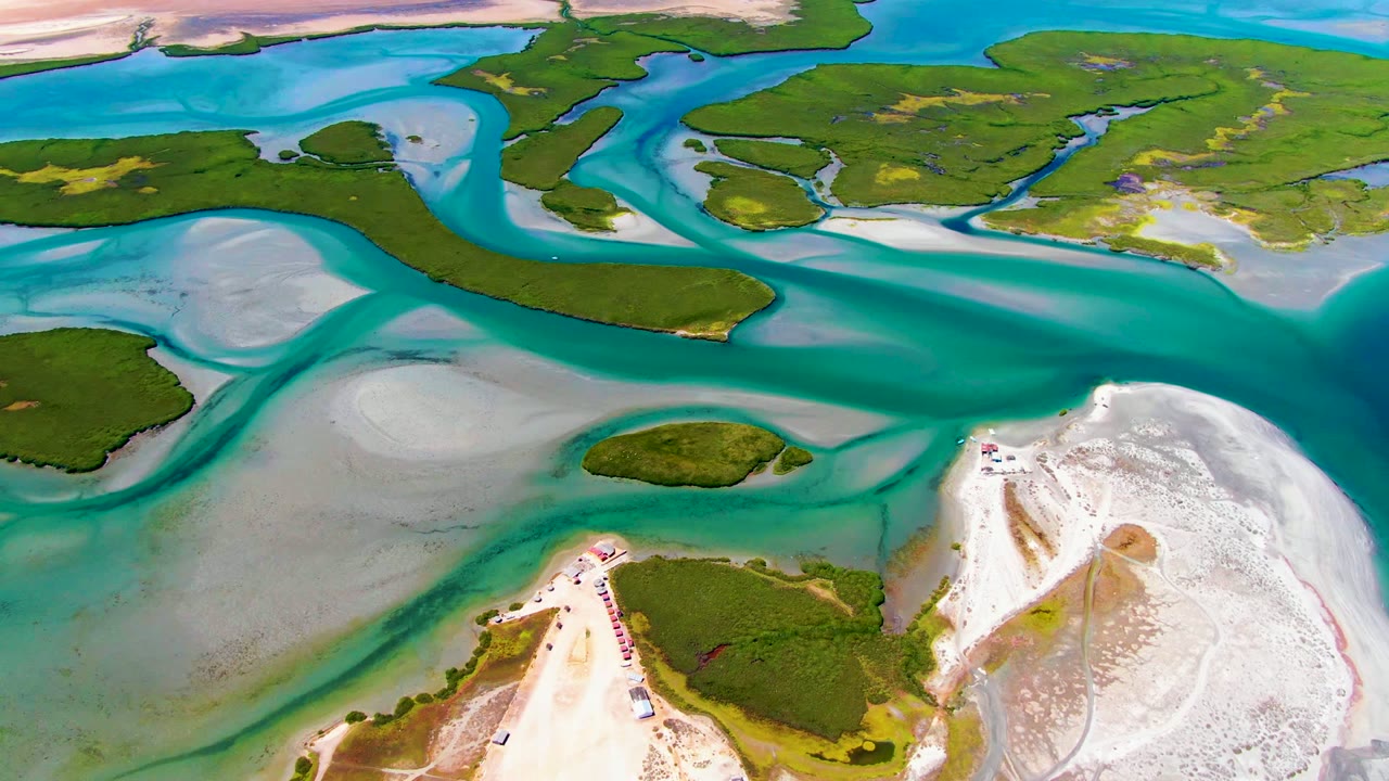 High aerial view over La Bocana, Baja California Sur, with turquoise water wrapping around green islands, pale sand flats, and a white beach point, with no visible people.