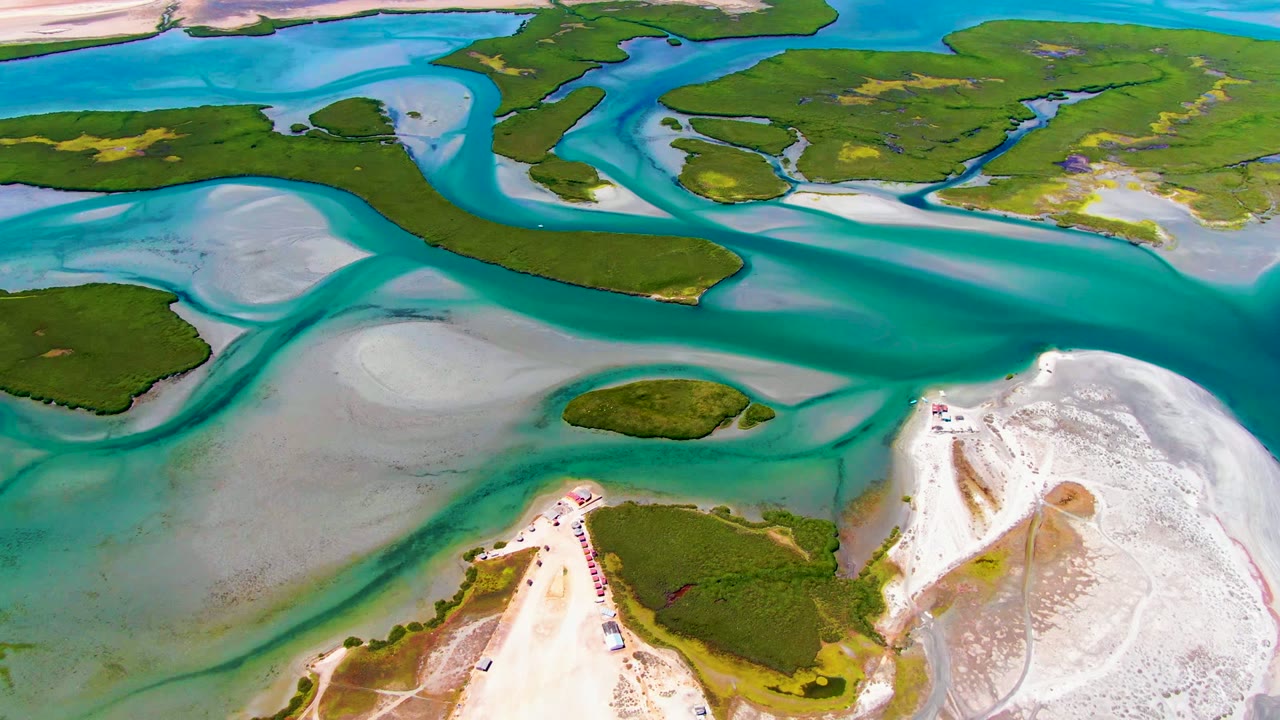 Aerial frame from La Bocana with a strong turquoise channel crossing the center, green island edges above and below it, and light sand flats along the shore, with no visible people.