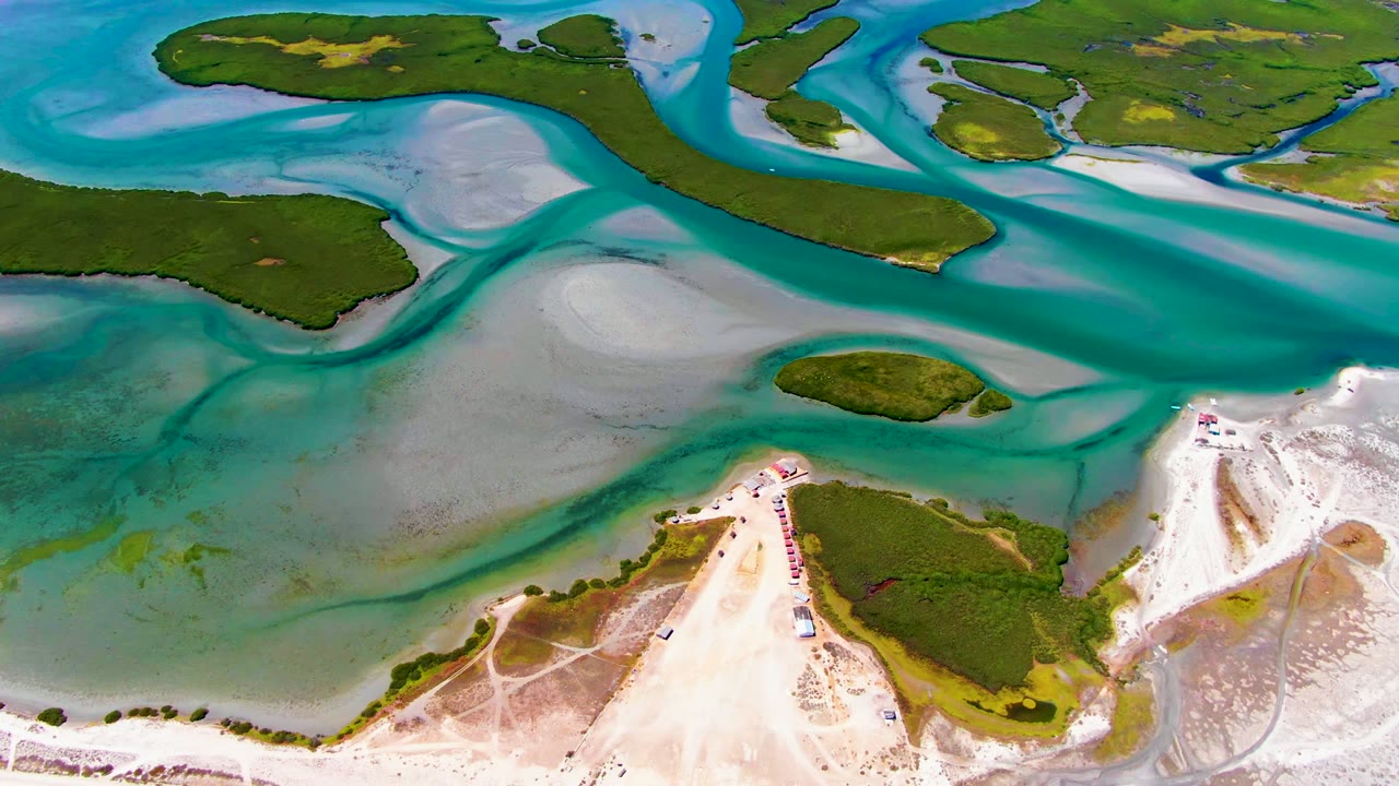 High aerial view over La Bocana with a teal main channel crossing the upper frame, muted sand flats below it, green islands on both sides, and no visible people.