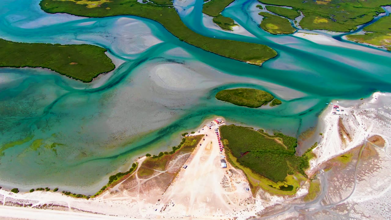Opening aerial frame over La Bocana, Baja California Sur, showing turquoise tidal water, pale sandbars, green islets, and no visible people.