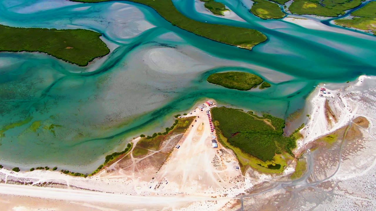 At 0:00, an overhead drone view in La Bocana shows turquoise tidal water wrapping around pale sandbars and small green islands, with no visible people.