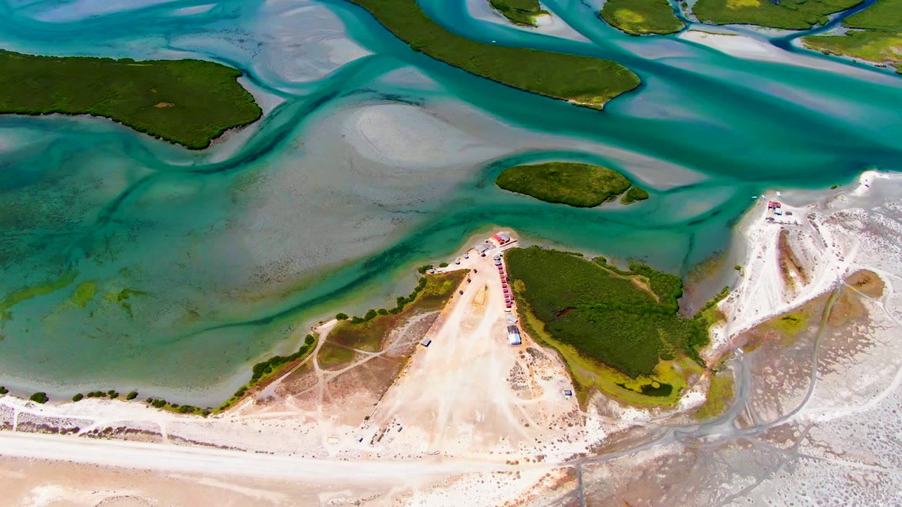 At 0:02, shallow turquoise water forms curved channels through exposed sand near small green landmasses in La Bocana, with no visible people.