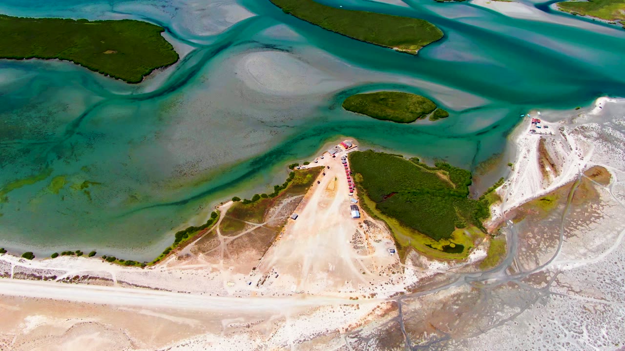 Overhead aerial frame of La Bocana with turquoise water channels, pale sand dunes, a beach strip, small green islands, and tiny beach activity near the sand point.