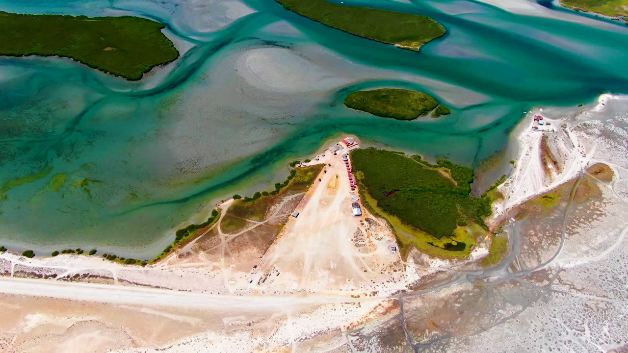 Top-down coastal lagoon with sand point, green island patches, and beach activity.
