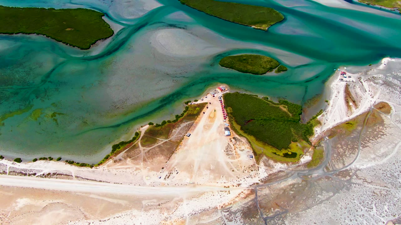 Overhead La Bocana frame showing turquoise water and darker channels above a pale shoreline, with dunes and small human activity clustered near the beach access area.