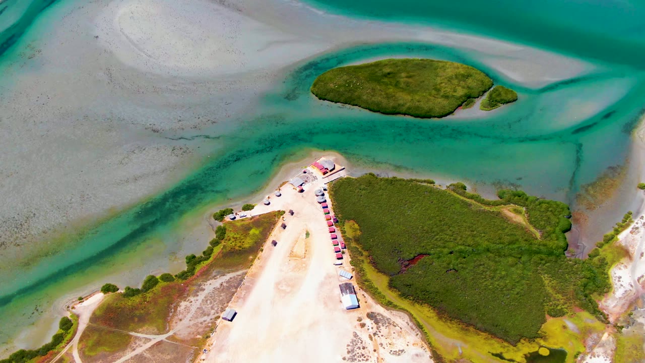 Aerial frame over La Bocana, Baja California Sur showing a pale sandy path, parked small vehicles, green shoreline vegetation, turquoise lagoon channels, and no visible people.