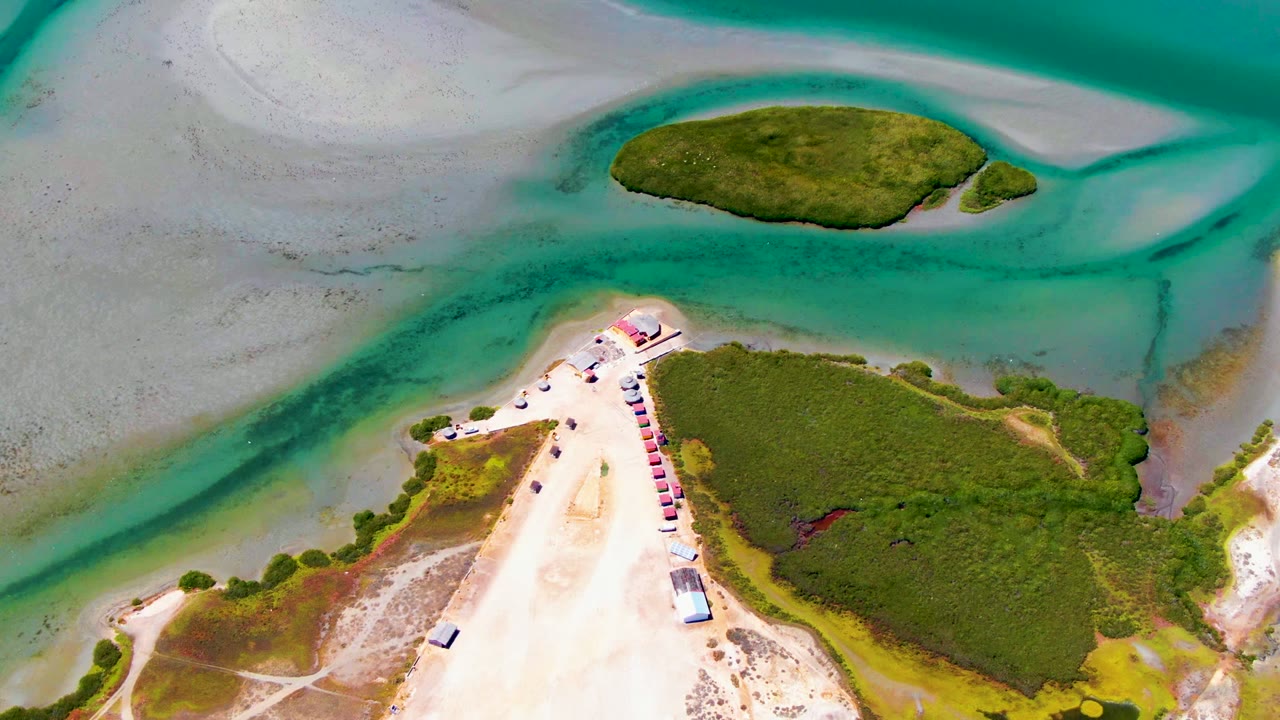 Overhead coastal view with a sandy track, vehicles, vegetation, and turquoise channels.