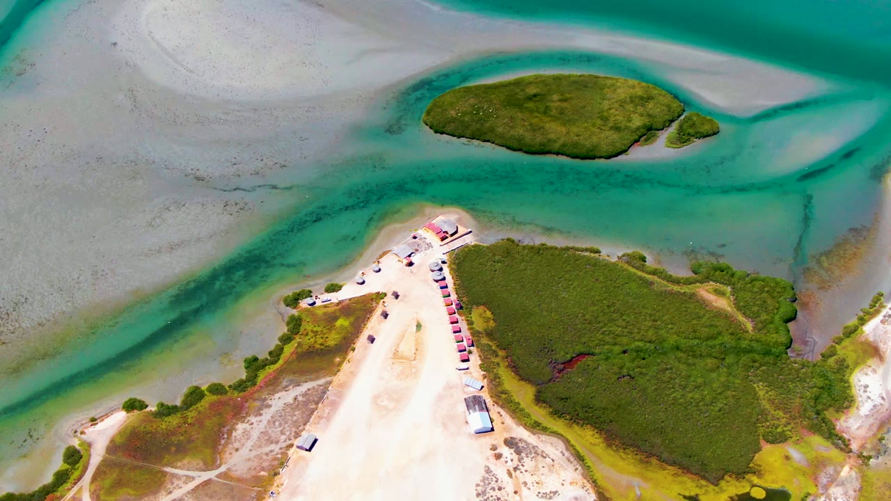 Mid-clip aerial frame in La Bocana with the pale sand path leading toward the water, green vegetation on both sides, a small offshore green landmass, and no visible people.