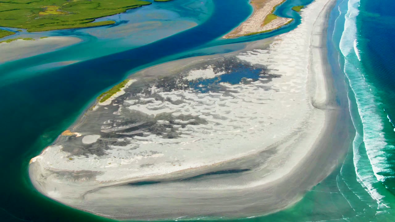 Aerial frame over La Bocana showing the sandbar shifted slightly across the frame, with turquoise water at the lower edge, a dark channel above, and a fresh band of surf foam at right with no visible people.