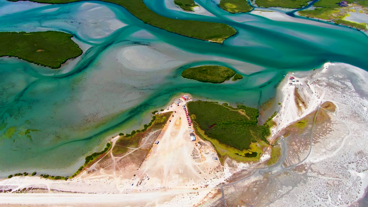 Overhead view of La Bocana tidal water around pale sandbars, green island patches, and a sandy shore with tracks and no visible people at the start of the clip.