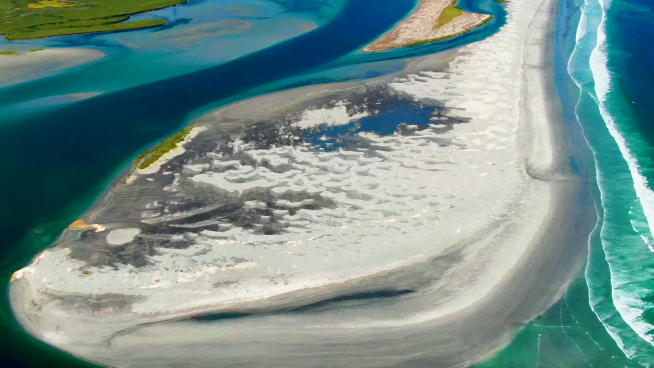 Wide aerial view in La Bocana, Baja California Sur, with pale sand, dark mottled patches, blue lagoon water, ocean surf lines, and no visible people.