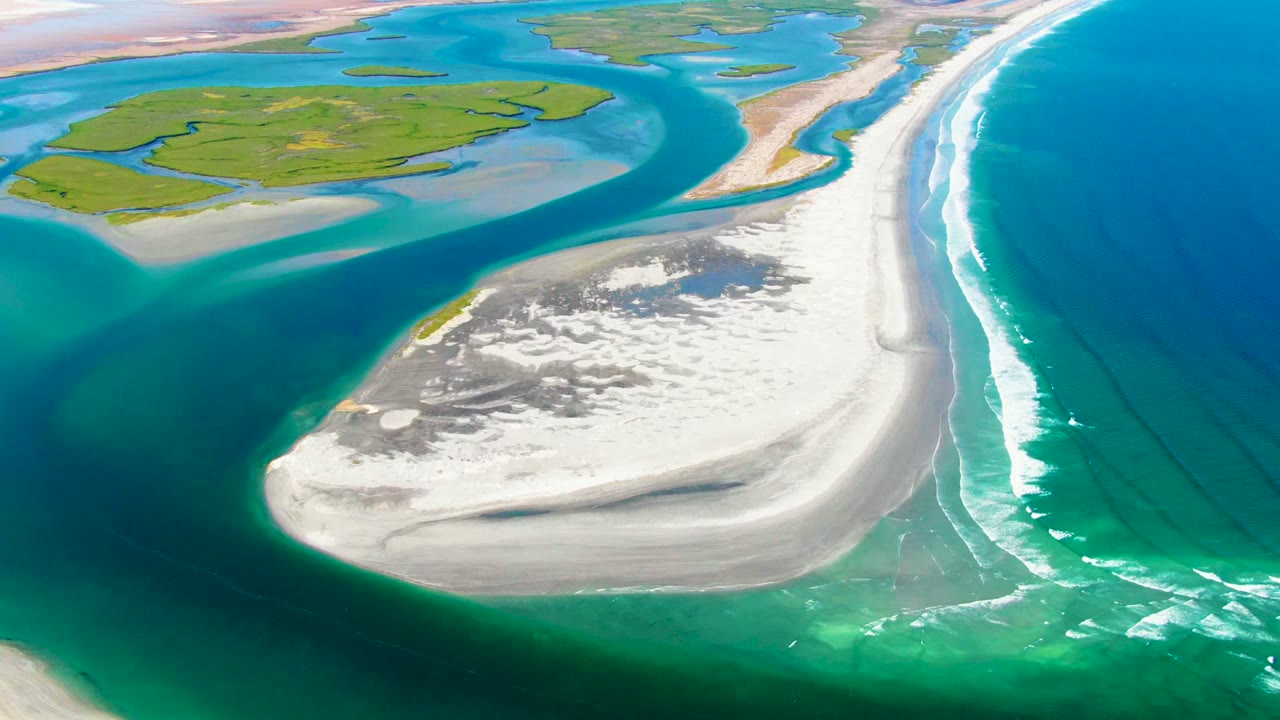 Aerial frame over La Bocana showing a broad white sandbar in the lower center, turquoise inlet water to the left, green land beyond, ocean surf at right, and no visible people.