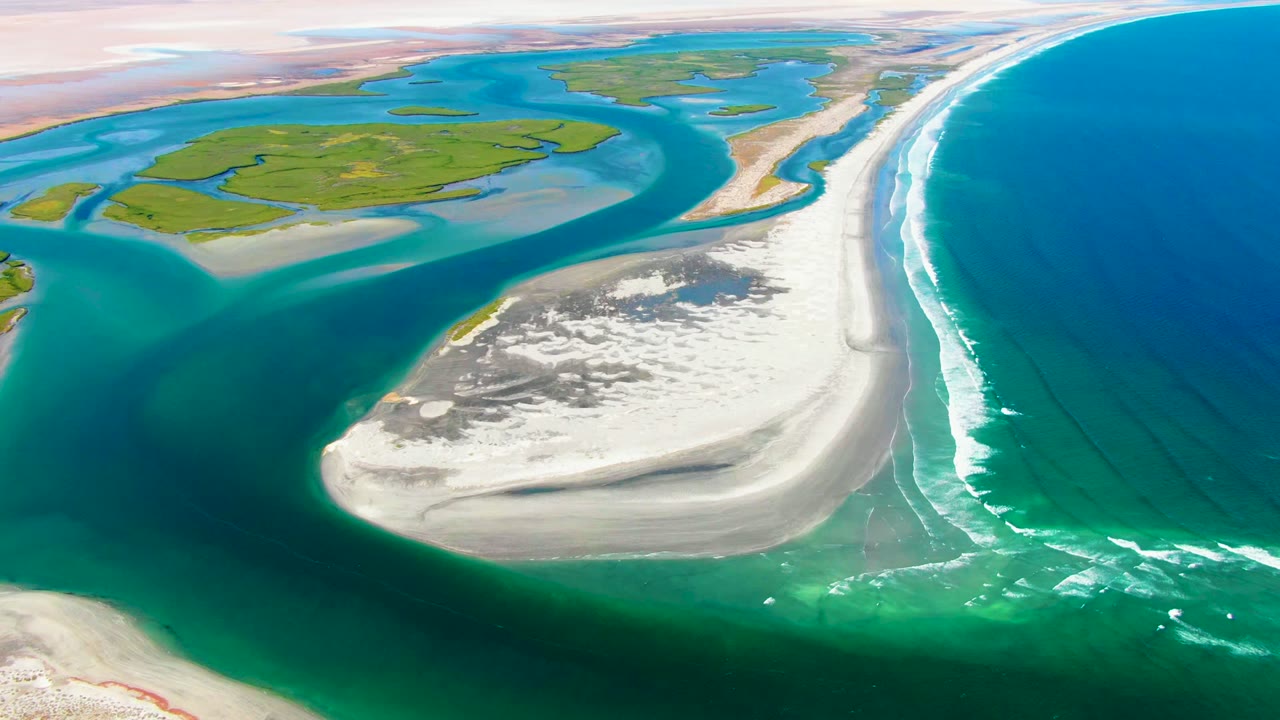 Aerial frame over La Bocana showing blue ocean filling the right side, a bright surf band beside the beach, pale sand in the lower middle, green lowland at top, and no visible people.