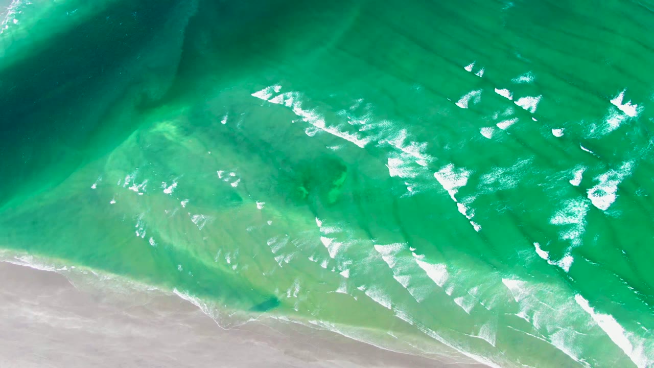 Overhead view of turquoise water and white foam reaching pale sand at La Bocana, Baja California Sur, with no visible people.