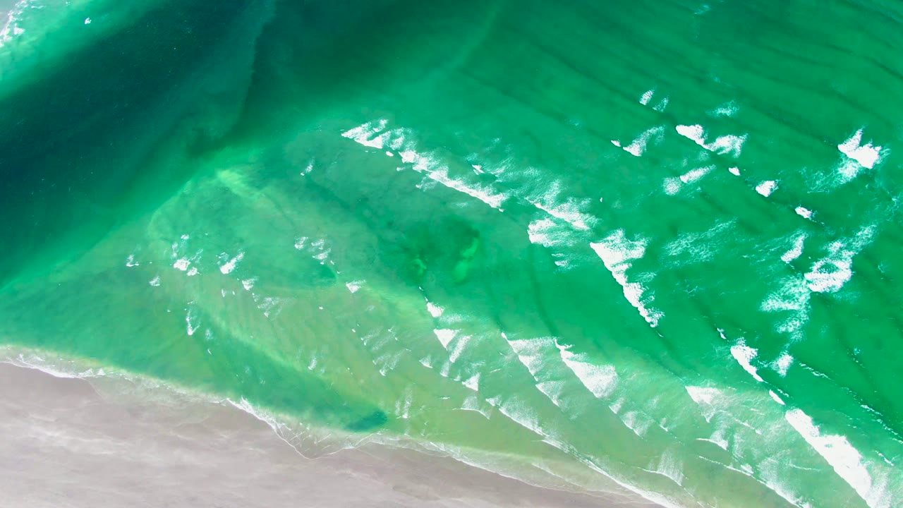 Aerial shoreline view with bright foam patches drifting through turquoise surf over pale sand in La Bocana, no visible people.