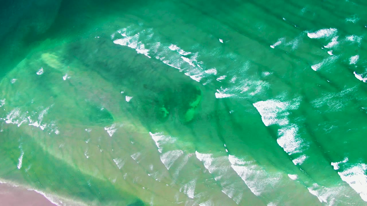 Straight-down aerial view of clear green ocean water over a pale seabed near La Bocana, Baja California Sur, with white foam bands entering from the upper right and no visible people.