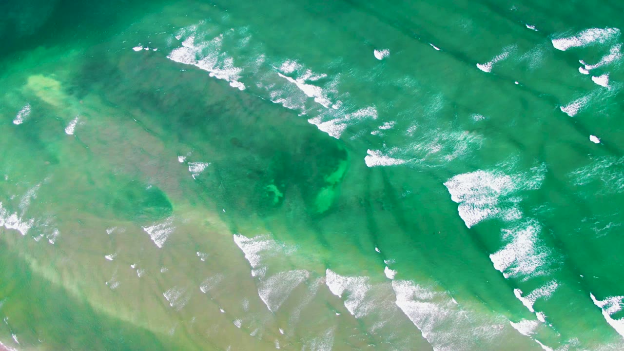Straight-down view of transparent green water near La Bocana, Baja California Sur, with broken white foam lines across the upper half and muted seabed forms below, with no visible people.