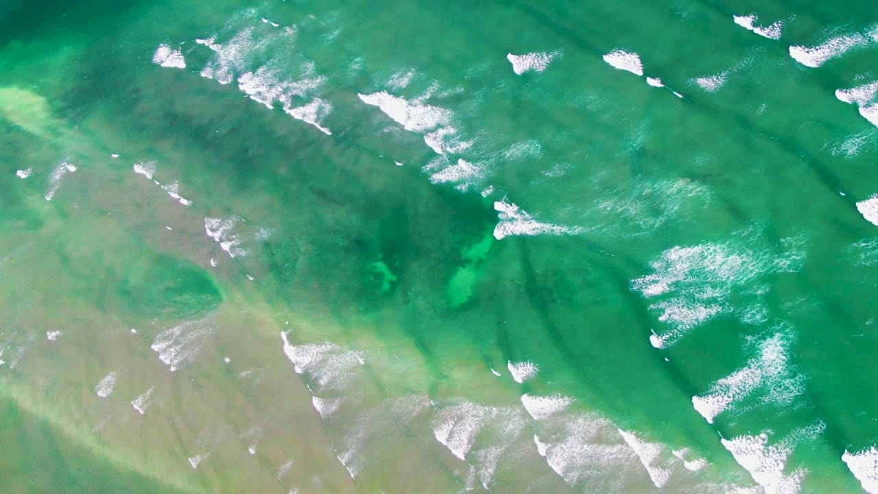 Aerial frame over green ocean water at La Bocana, Baja California Sur, with scattered white foam near the upper edge and no visible people.