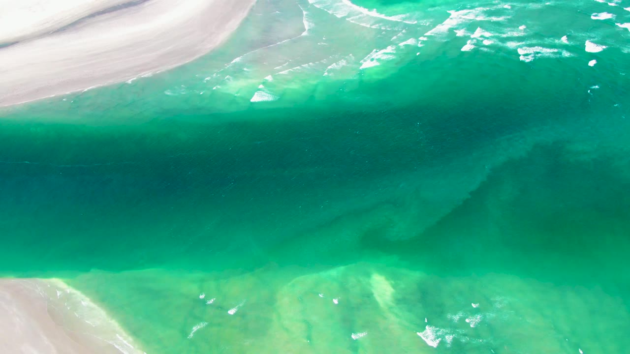Aerial view of La Bocana shoreline showing turquoise water, pale sand entering from the upper and lower edges, small foam marks, and no visible people.