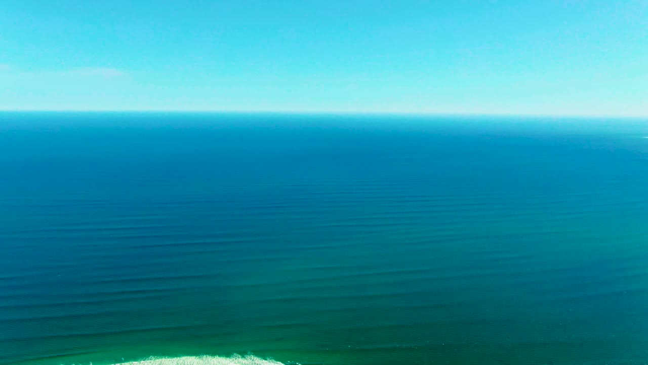 Aerial view over La Bocana ocean water with the horizon across the top, turquoise shallows below, and a small white break appearing at the lower right with no visible people.