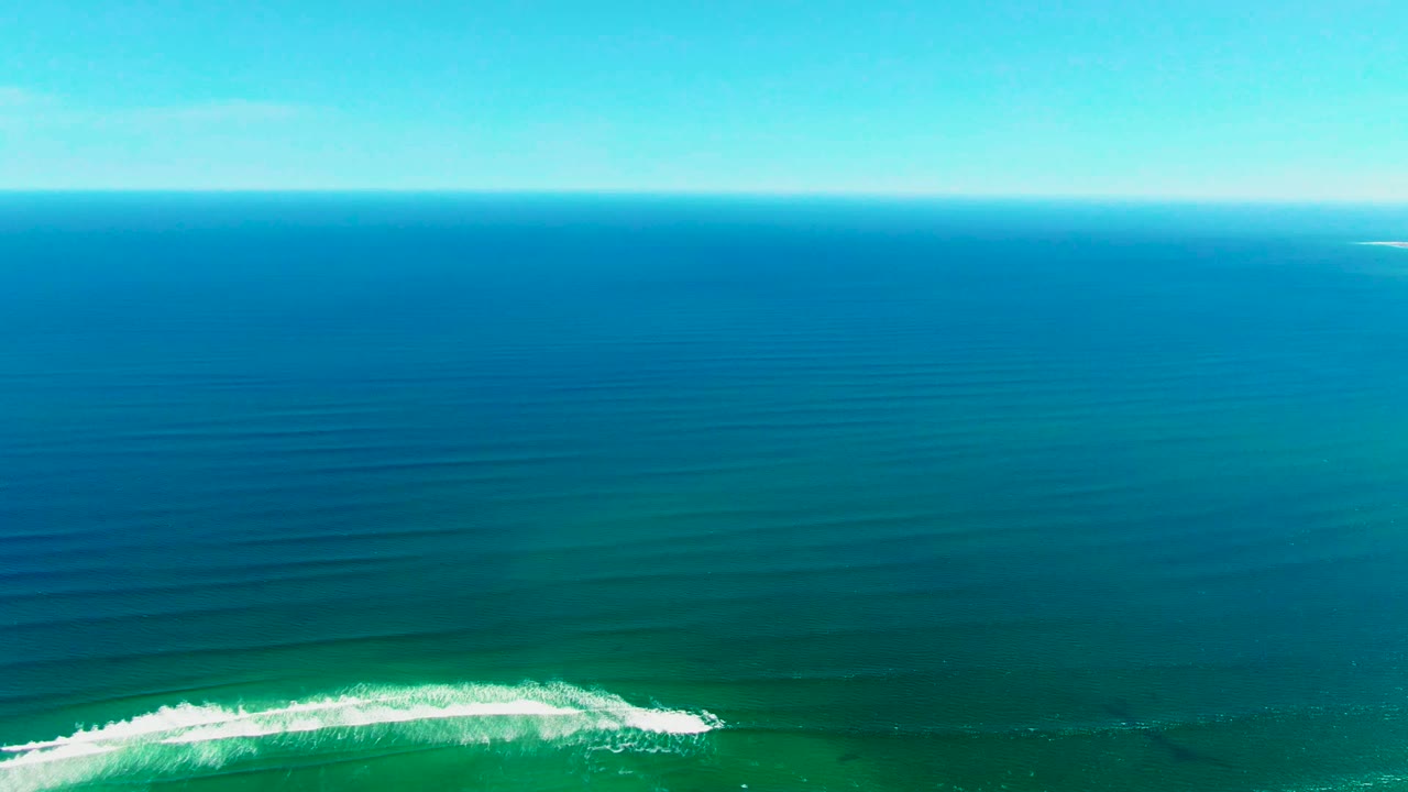 Aerial view over La Bocana ocean water as a white wave begins breaking along the lower edge, with deep blue sea and clear sky above, no visible people.