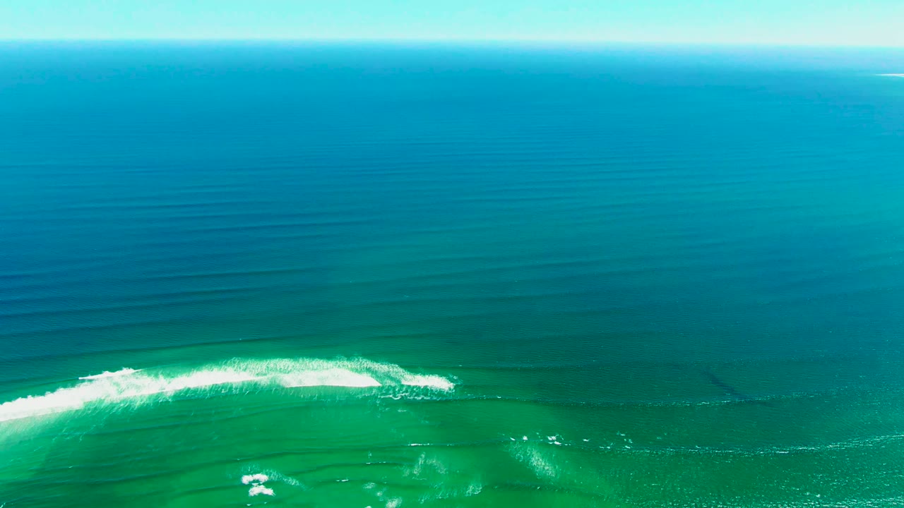 First frame of an overhead ocean view in La Bocana, Baja California Sur, with turquoise water, darker blue bands, a white breaking wave low in frame, and no visible people.