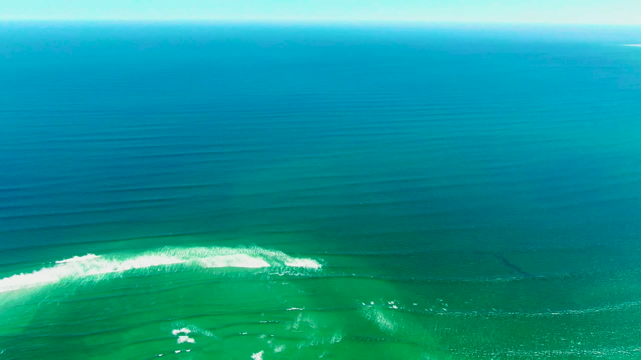 Aerial ocean surface with a white wave crest spreading across turquoise water.