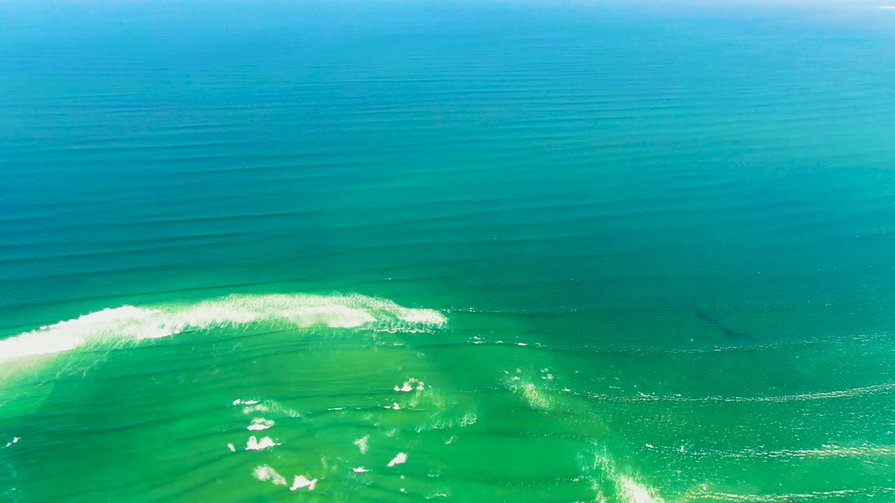 Aerial view over clear water at La Bocana, Baja California Sur, with deep blue sea above, green shallows below, a white wave beginning to break, and no visible people.