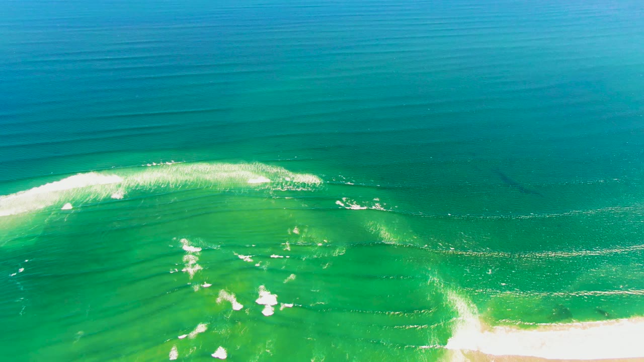 Aerial frame over La Bocana, Baja California Sur, with turquoise shallows, white foam spreading near the lower left, pale sand at the bottom edge, and no visible people.