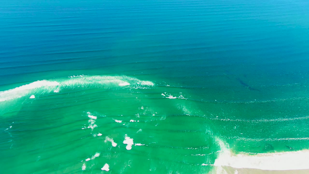White surf curls across turquoise water beside a sandy shore.