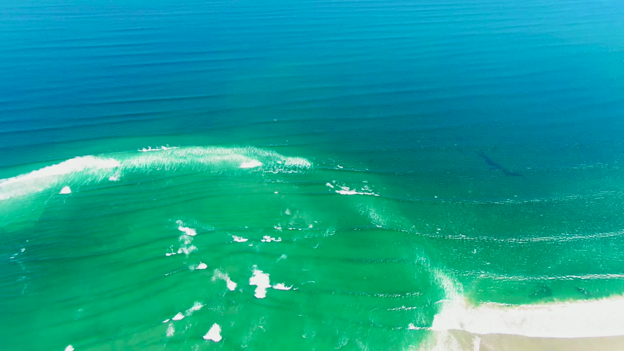Aerial frame over La Bocana with deep blue water above, a broad turquoise band below it, scattered white foam patches in the lower center, sand at the bottom right, and no visible people.