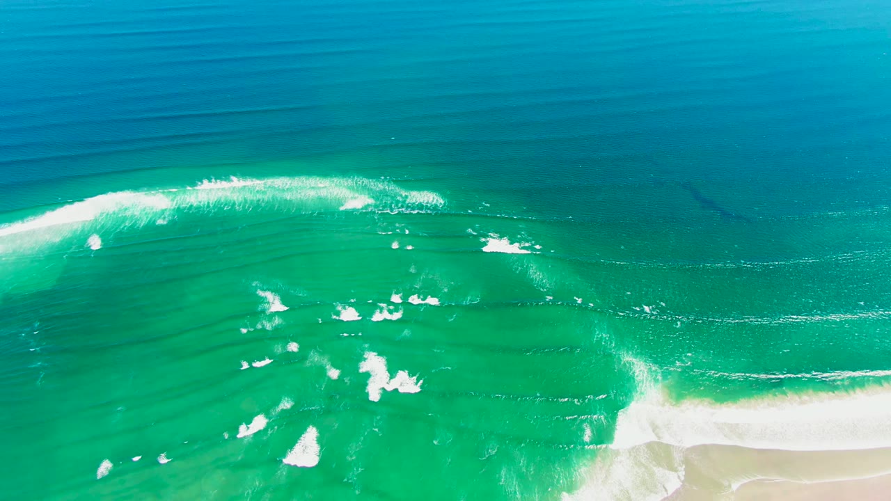 Aerial frame from La Bocana, Baja California Sur showing turquoise water, white foam patches, deep blue ocean beyond, pale sand at the lower right, and no visible people.